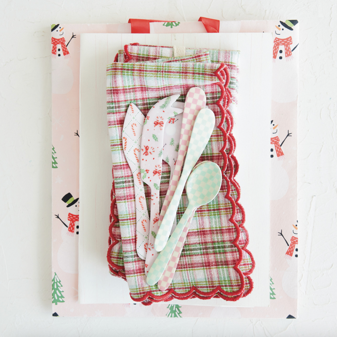 A set of Christmas-themed tableware and napkins featuring a festive red, green, and white plaid pattern, complete with snowman illustrations on the wrapping paper. The set includes a napkin with a red scalloped edge, matching patterned spoons, and a small checkered spoon, all arranged on a white background.