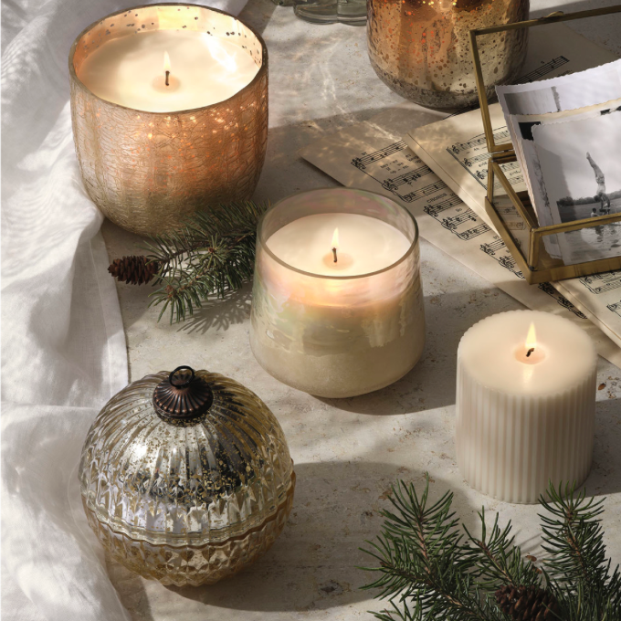 A styled arrangement of multiple glass and metallic candle holders with lit candles on a draped fabric surface, surrounded by pine branches, a small pinecone, and a gold-toned photo frame.