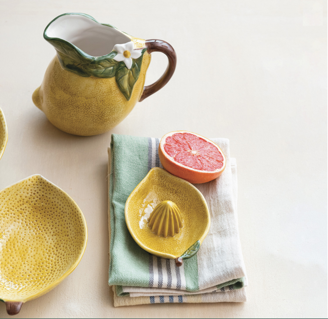 Yellow ceramic lemon-shaped pitcher, bowl, and citrus press on a light surface with a napkin and half a grapefruit.