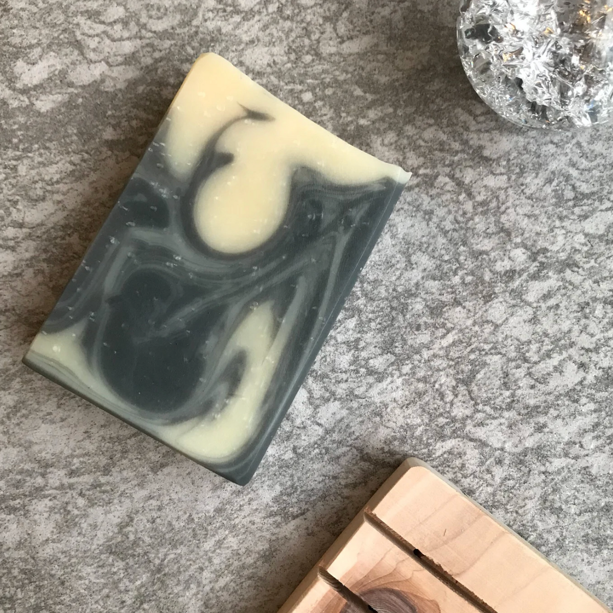 A rectangular bar of marbled soap with dark gray and creamy off-white swirls, placed diagonally on a textured gray stone surface next to a wooden block and a clear decorative crystal.