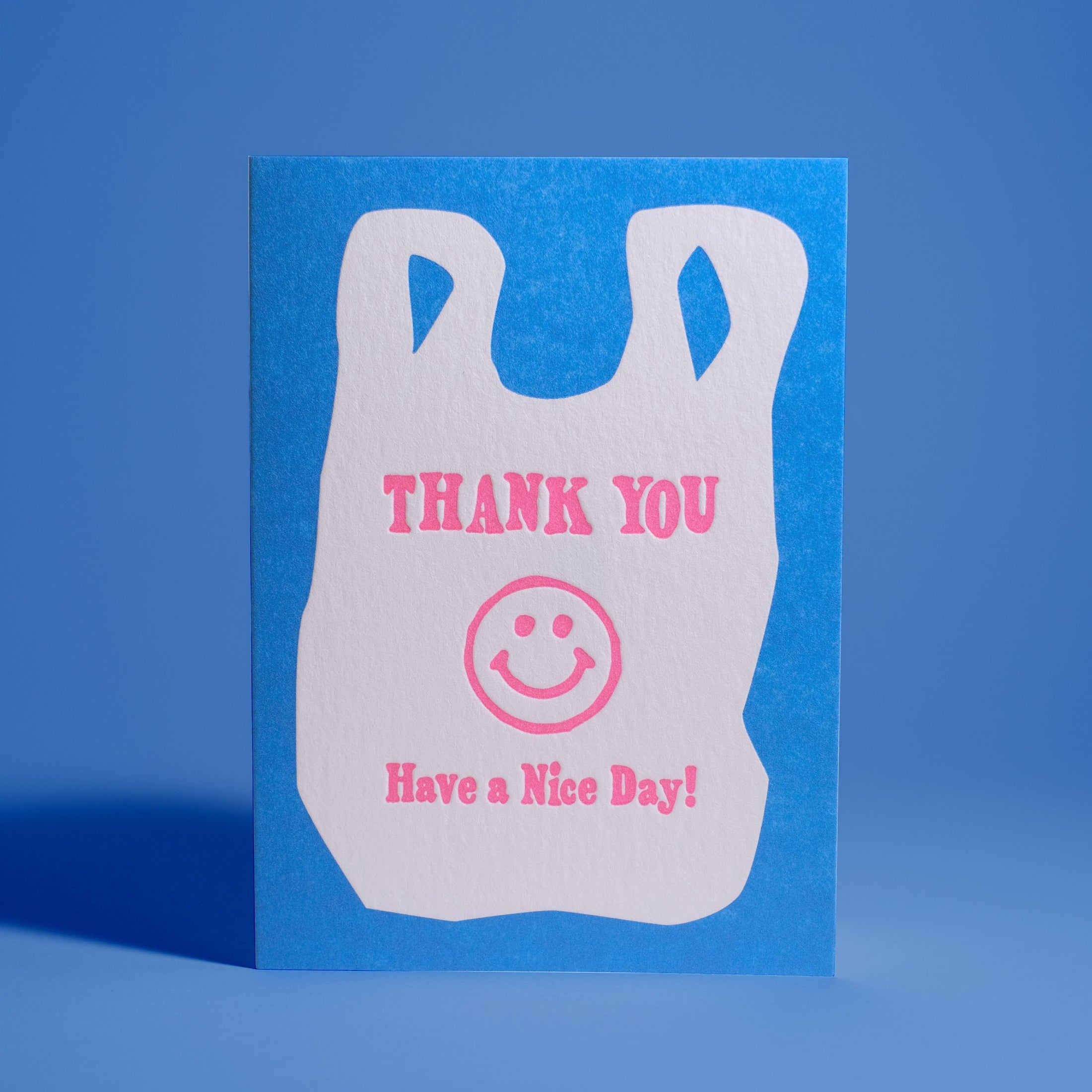 Bright blue greeting card with a white bag design that says "Thank you, have a nice day!" with a cute smiley face. Lettering done in pink. 