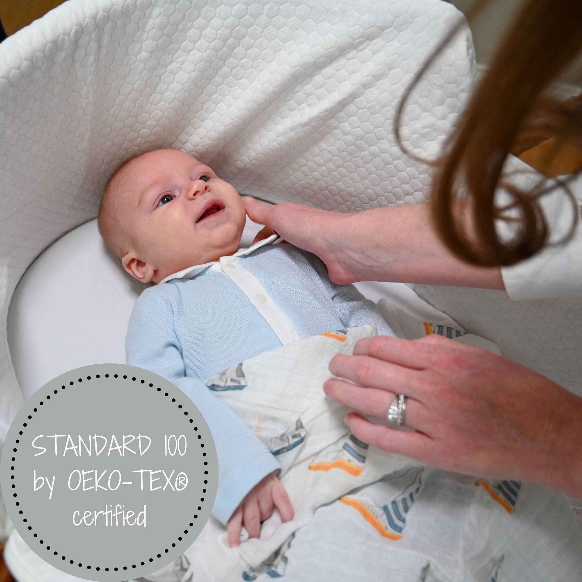 Baby lying in a crib with a person adjusting a blanket, featuring OEKO-TEX certification.