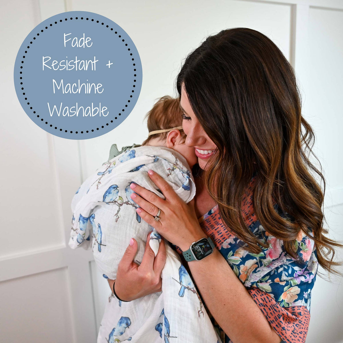 A woman holding a baby wrapped in a white muslin swaddle with a blue bird print. A blue circular badge in the top left reads 'Fade Resistant + Machine Washable'.