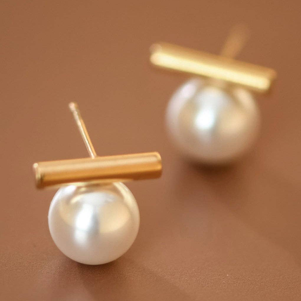 Elegant white pearl studs with a delicate gold bar accent, crafted from hypoallergenic stainless steel and finished with 18K gold plating. Measuring approximately 0.55" x 0.5", these minimalist earrings are waterproof and tarnish-resistant, making them perfect for everyday wear. To maintain their lustrous finish, avoid prolonged contact with lotions, perfumes, and cleaning products. A timeless addition to any jewelry collection.