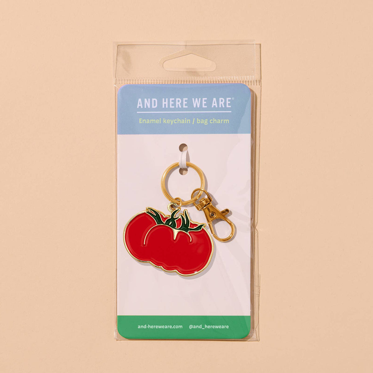 Red tomato-shaped keychain on a packaging card with 'And Here We Are' branding on a beige background.