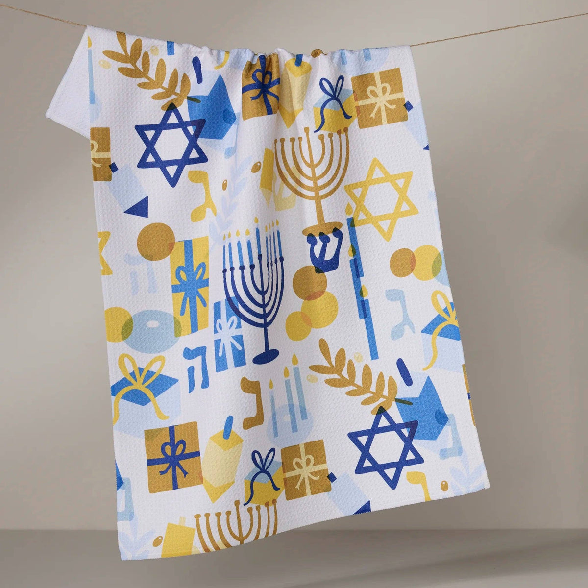 A white kitchen towel with a colorful Jewish holiday pattern featuring blue and yellow Star of David, Menorah, and gift boxes, hanging from a string against a neutral beige background.