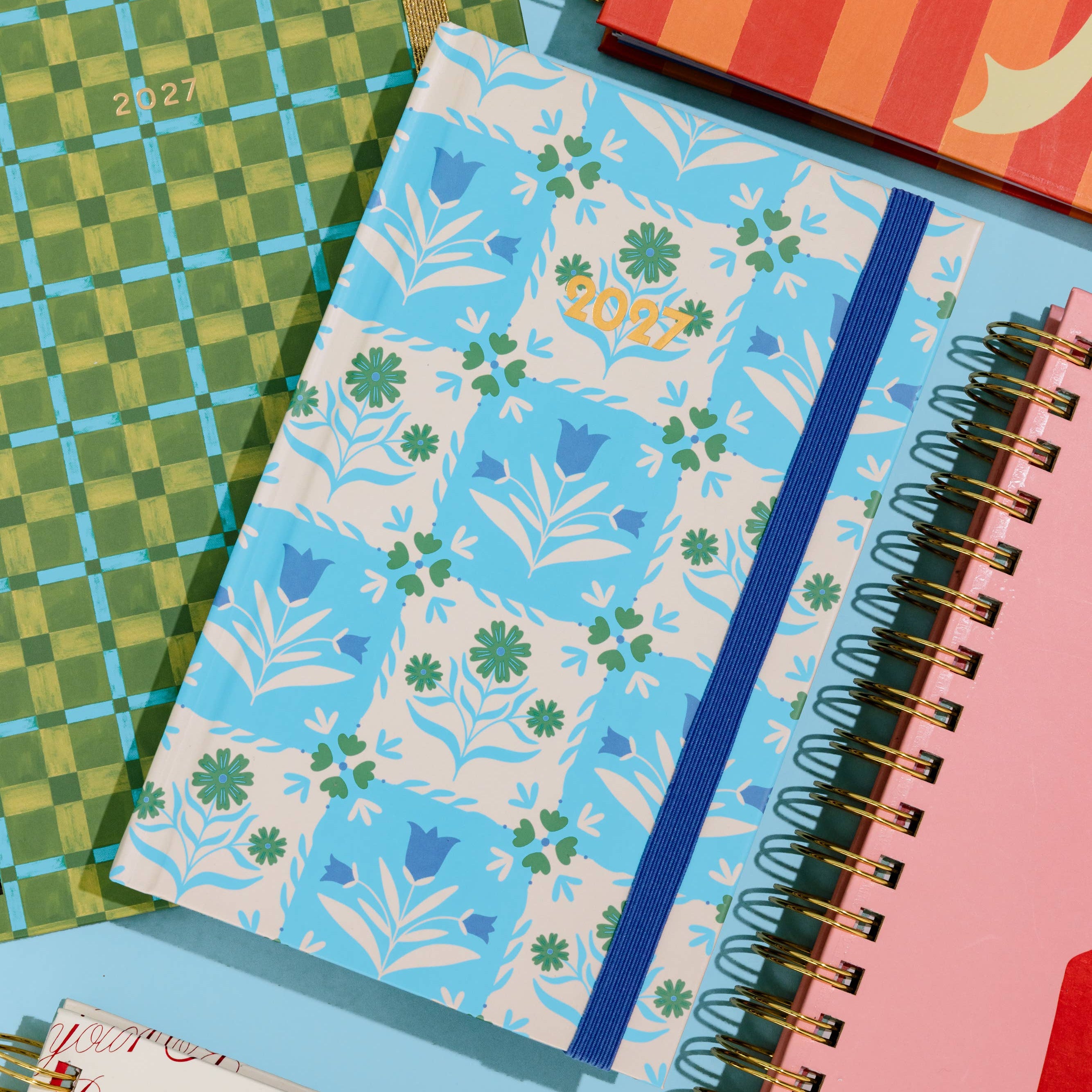 A styled flat-lay of several colorful spiral notebooks and journals on a light blue background. The central notebook has a light blue and white floral patchwork pattern with a dark blue elastic band.