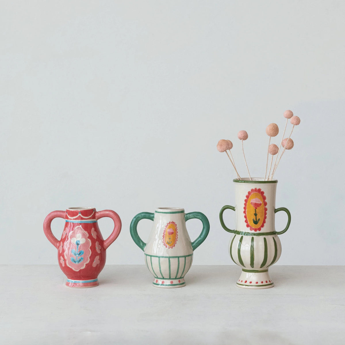This Hand-Painted Stoneware Urn with Flower, Stripes, and Handles is a celebration of artistry and color. Its multi-colored palette includes a range of hues that complement its unique design. The urn's other shape sets it apart, offering a fresh take on traditional vases. Influenced by Scandinavian, Boho, and French Country styles, it serves as a versatile decor piece