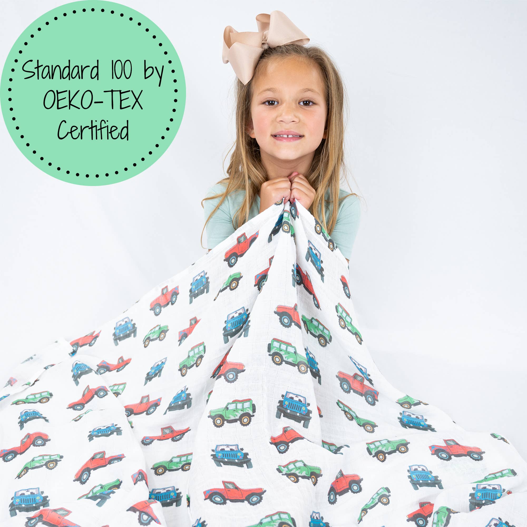 Child holding a colorful blanket with vehicle patterns, OEKO-TEX Certified label on the side.
