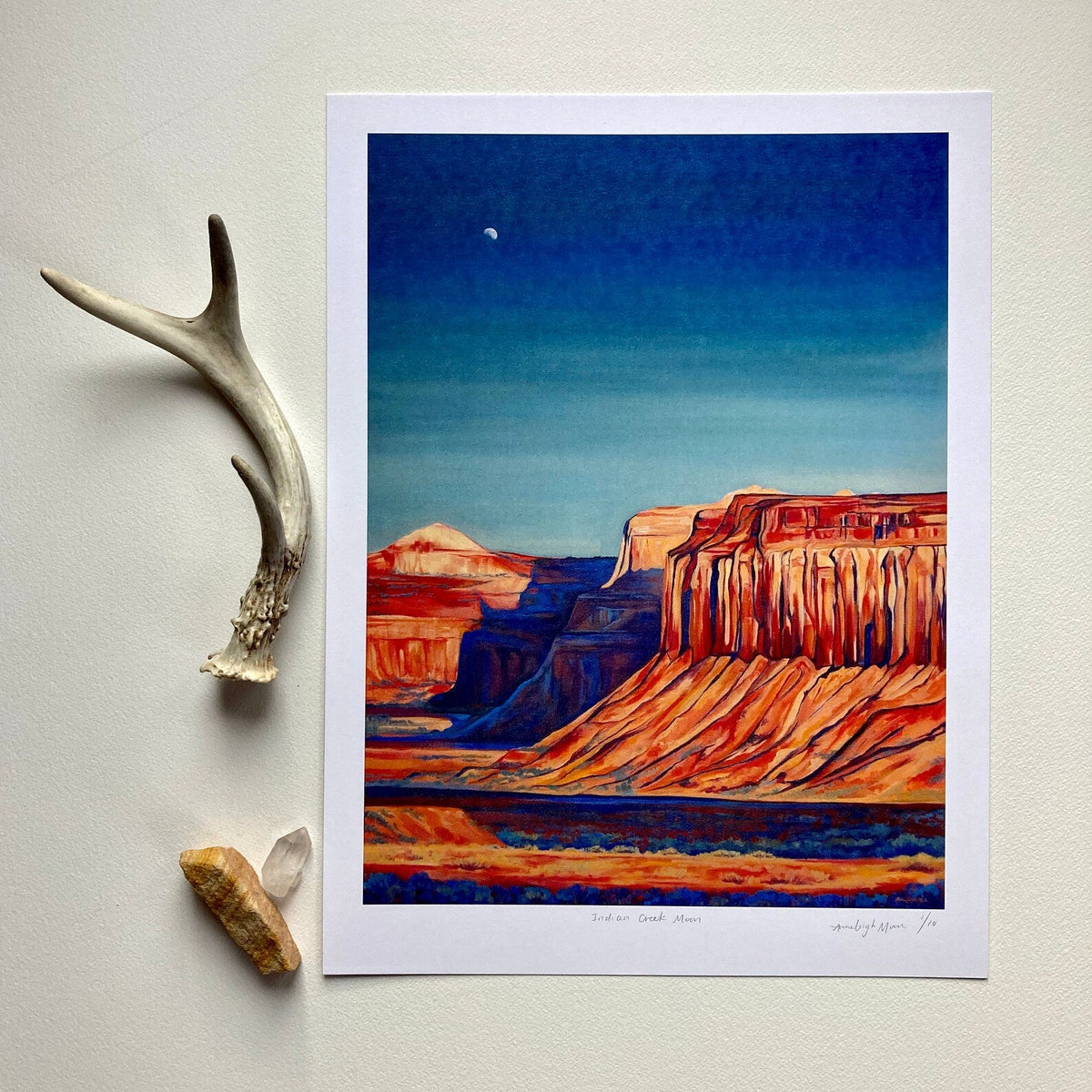 Art print of a desert landscape with a blue sky, placed on a light surface with a deer antler and crystal beside it.