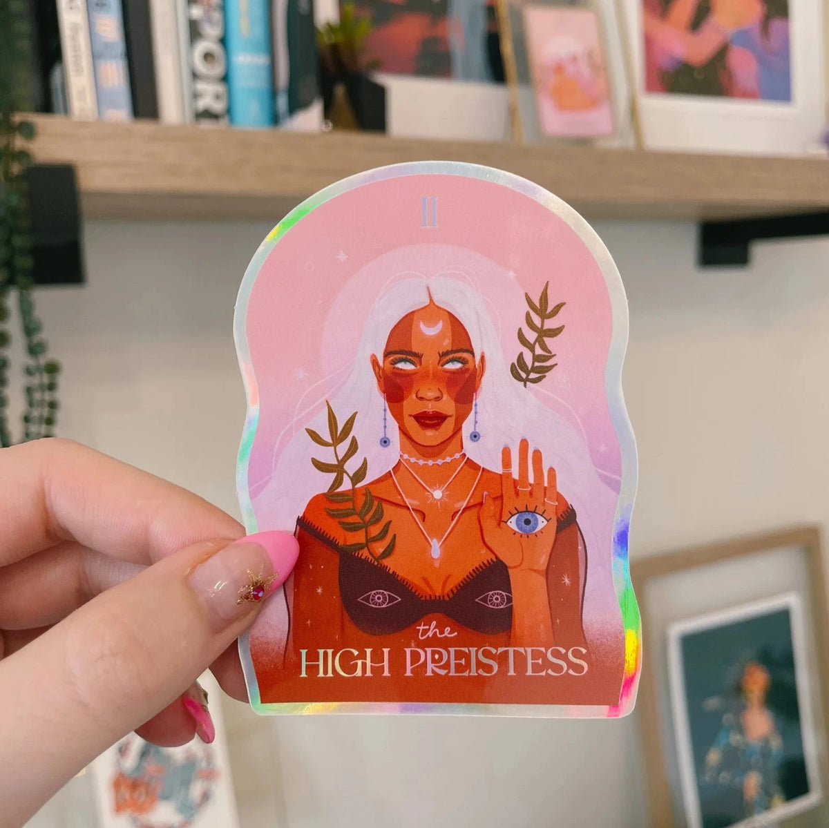 The High Priestess Sticker with holographic effects. The high priestess is holding her left hand up and in her palm is an eye. She has a bra on with an eye on each cup and she is wearing sparkly silver necklaces. She has long, wavy white hair. 