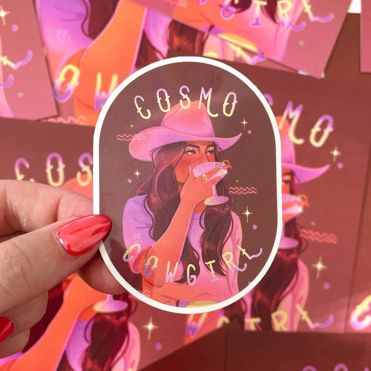 Cosmo Cowgirl sticker with mirrored foil effects. The cowgirl is wearing a cowgirl hat and plain t shirt and is drinking a cosmo out of a glass coup. She has long brunette hair and the sticker is in a hue of pink and purple. 