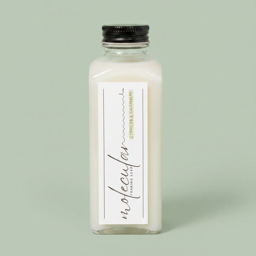 A hand holds a clear rectangular bottle of Molecular Foaming Soap  labeled Cypress and Cashmere. The bottle contains a pale liquid and is photographed against a soft green background
