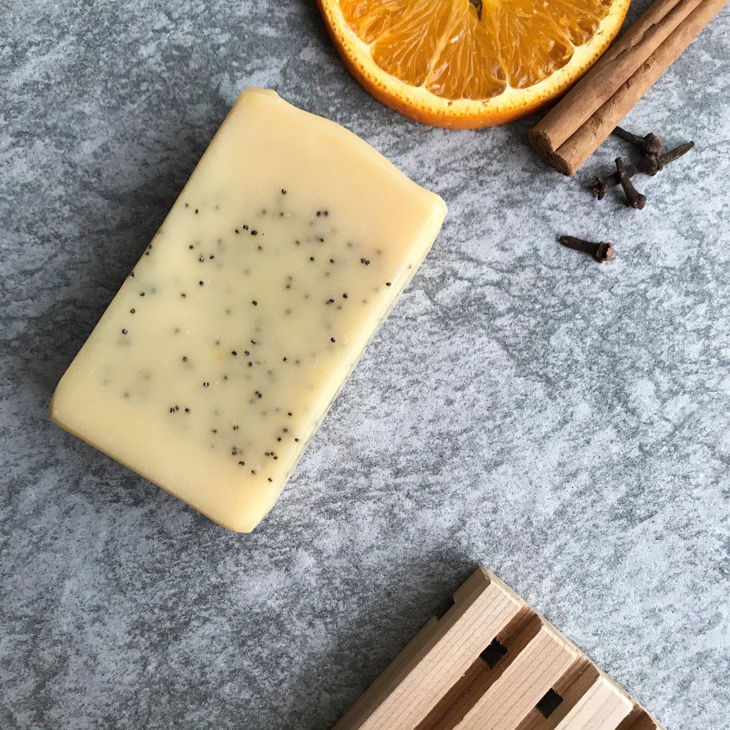 Bar of soap with specks on a gray surface with orange slice, cinnamon stick, and cloves.