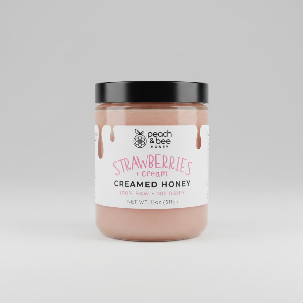 A jar of Peach & Bee creamed honey with strawberries and cream flavor, placed on a table with whole strawberries and a bowl in the background.