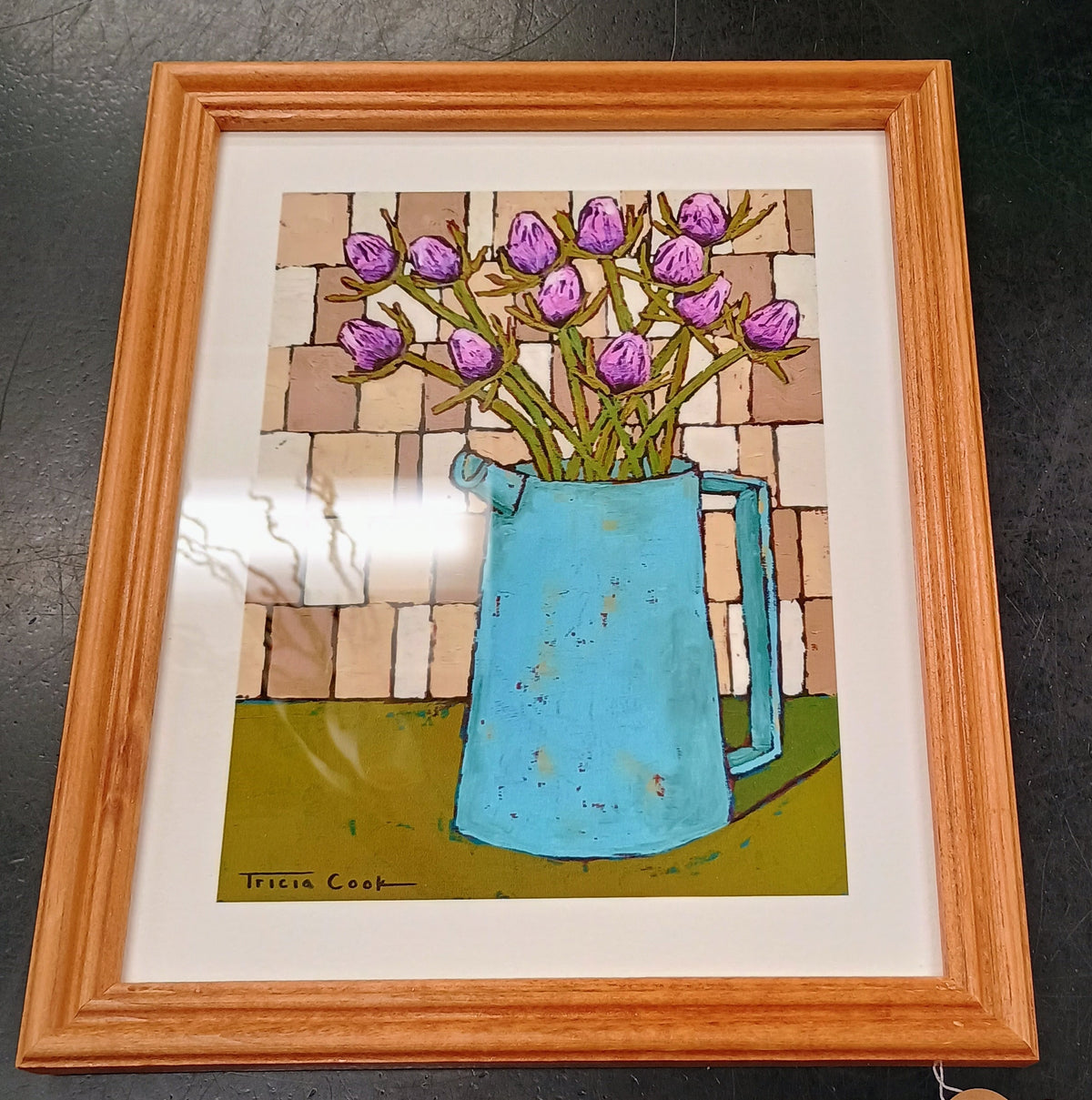 Framed print of purple thistle buds in a sky blue pitcher, on a green table top, against a light brown tile wall.  Frame is oak